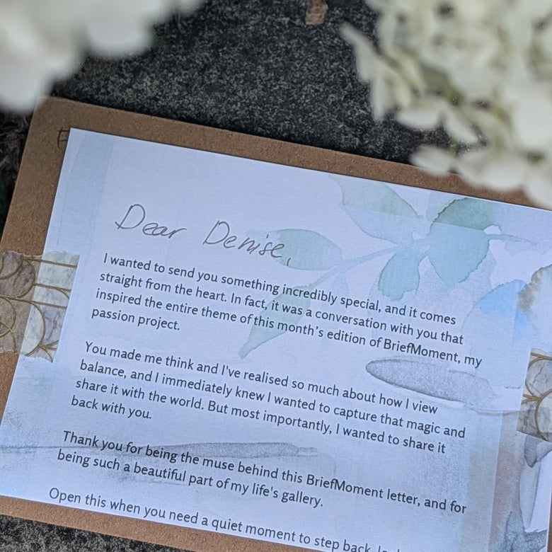 A close-up of a printed briefmoment letter  placed on a brown kraft envelope, with soft white flowers blurred in the foreground and a dark textured surface underneath.