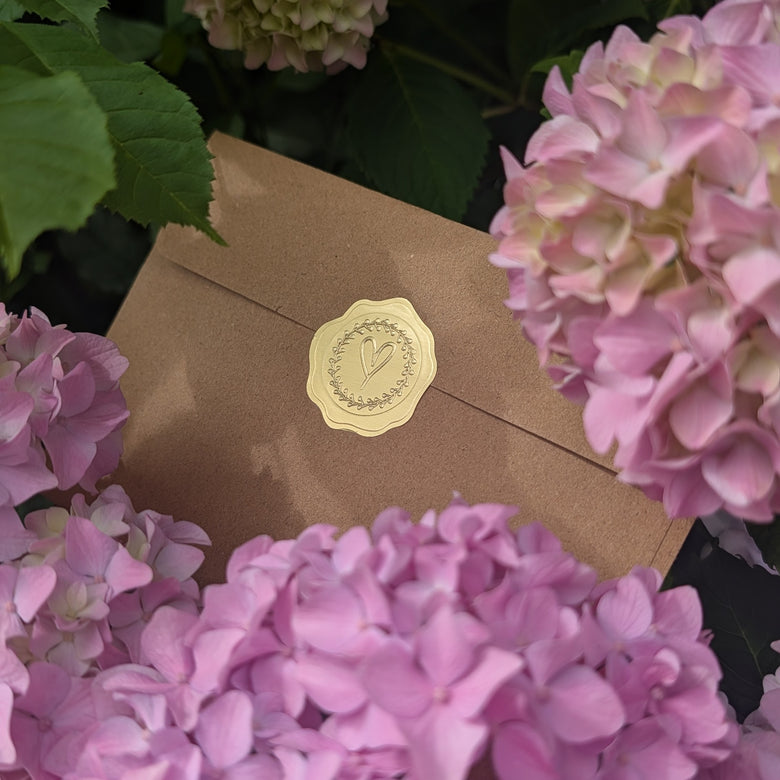 A kraft brown briefmoment envelope with a round gold seal featuring an embossed monogram, nestled among pink hydrangea flowers and green leaves.