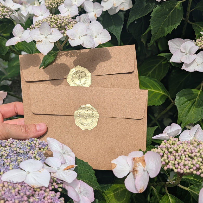 Two kraft paper envelopes from briefmoment with gold seals are held by a hand against a background of green leaves and white and purple hydrangea flowers.