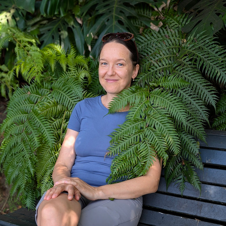The certified coach named Ksenia Kram with sunglasses on her head sits on a dark wooden bench surrounded by lush green ferns and tropical plants. She is wearing a blue short-sleeve t-shirt and gray pants.