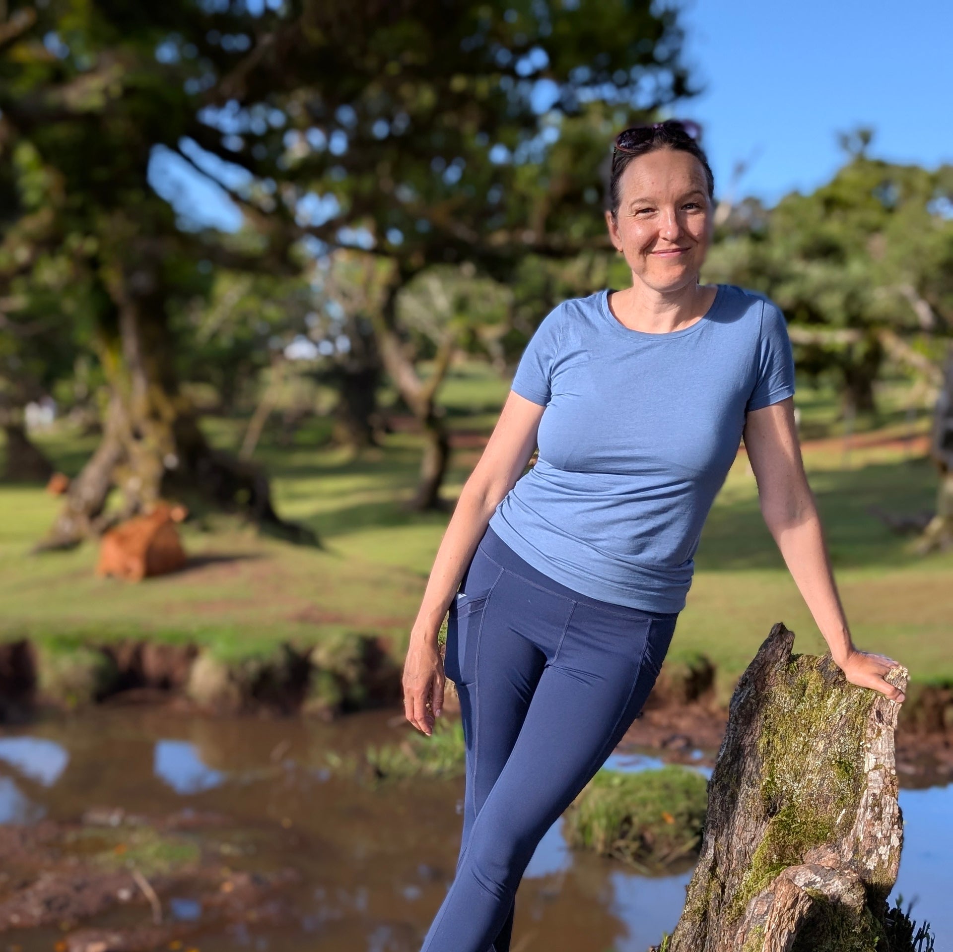 The certified coach named Ksenia Kram wearing a light blue short-sleeve t-shirt and navy blue leggings stands outdoors next to a log in a park-like setting with trees and water in the background.