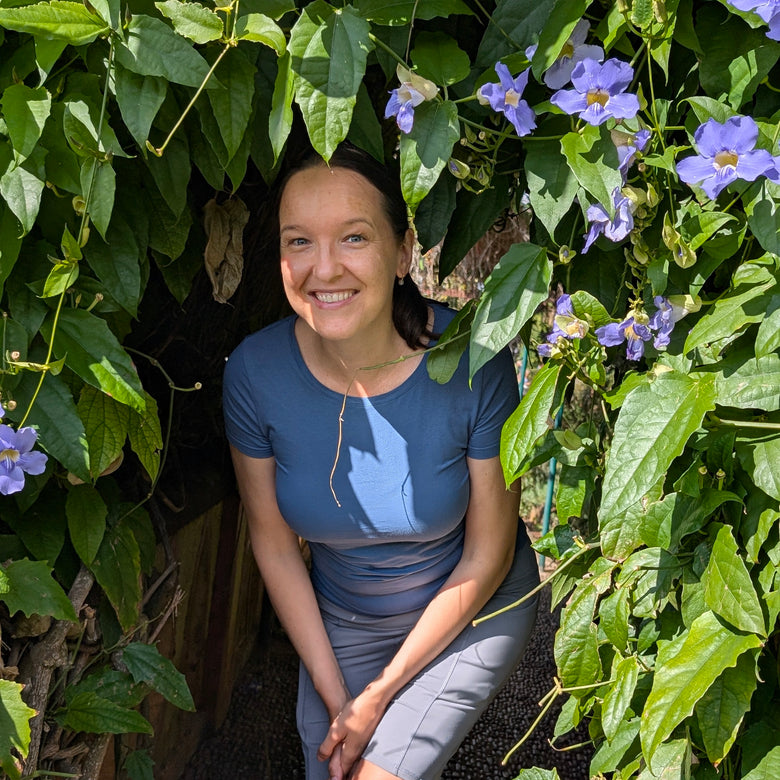The certified coach named Ksenia Kram smiling while wearing a slate blue short-sleeve athletic top and gray leggings, posing outdoors among lush green vines and purple flowers.